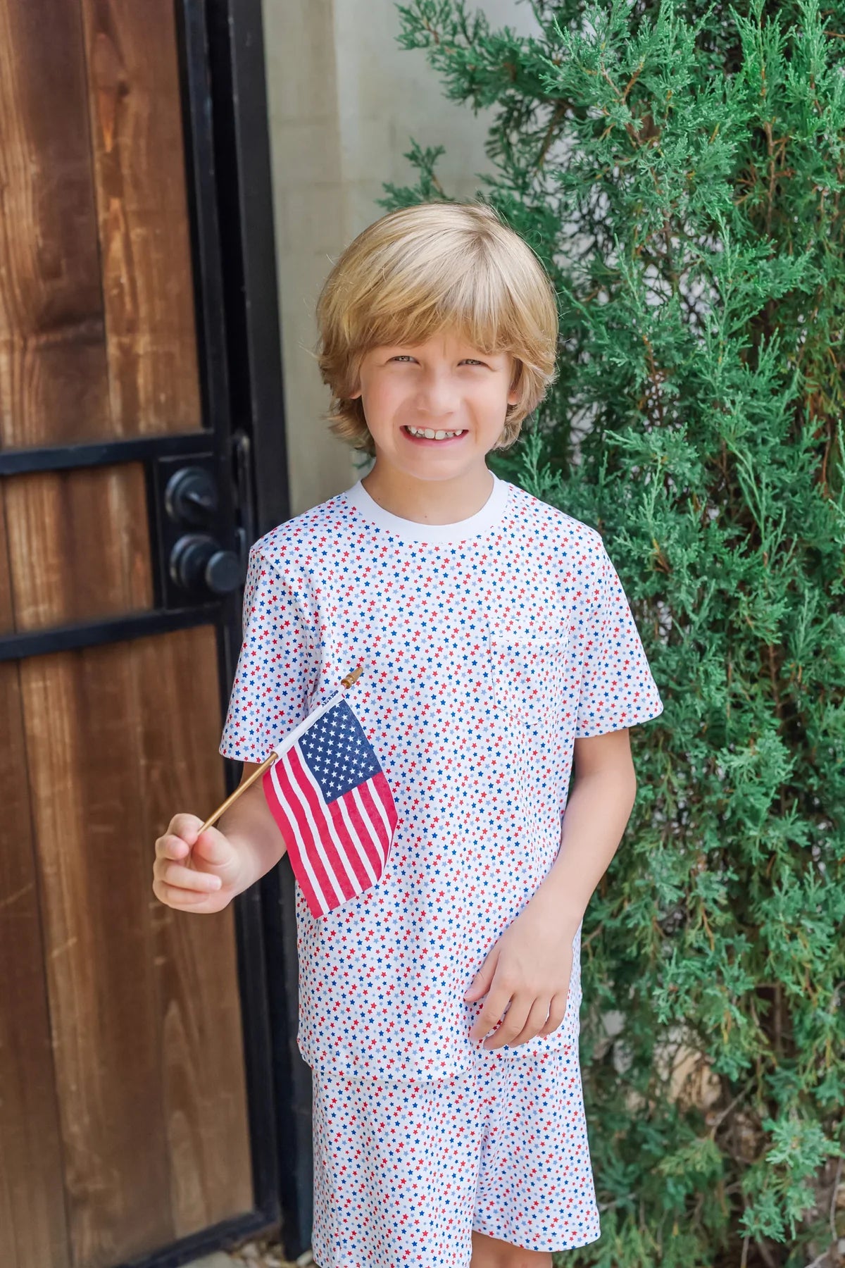 A smiling child in the Lila & Hayes Boys' Charles Short Set holds a small American flag while standing outside by a wooden door and green shrub.
