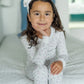 A young girl with long brown hair and a white headband smiles softly while sitting on a neatly made bed, dressed in the snug-fit Lila & Hayes Baby Girls' and Girls' Ava Pajama Set.