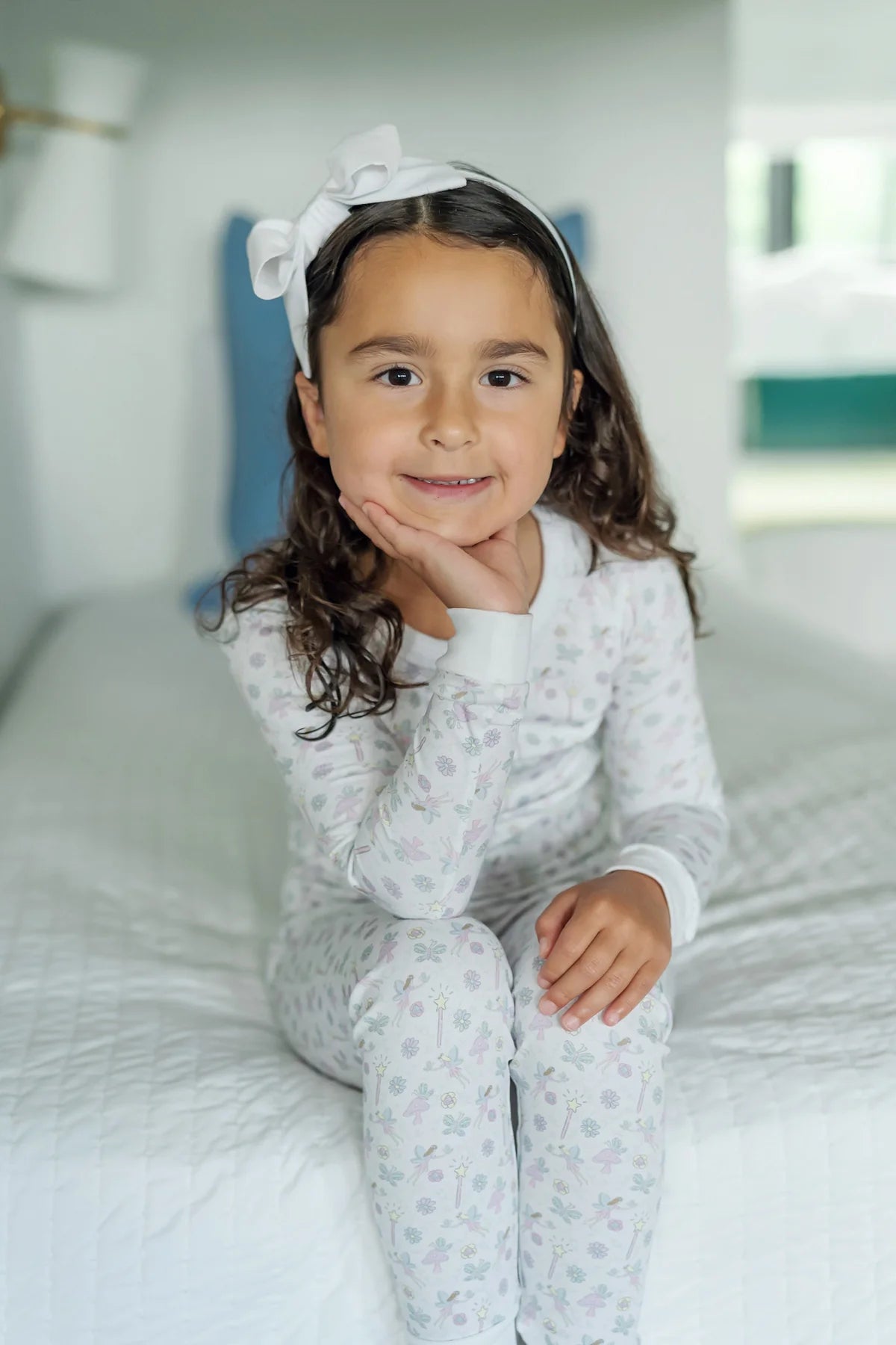 A young girl with long brown hair and a white headband smiles softly while sitting on a neatly made bed, dressed in the snug-fit Lila & Hayes Baby Girls' and Girls' Ava Pajama Set.
