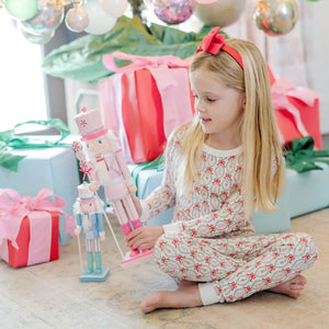 A young girl in the Lila & Hayes Girls' Ava Pajama Pant Set sits on the floor holding a pink nutcracker, surrounded by pastel-wrapped gifts and festive Christmas decorations.