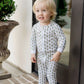 A toddler stands barefoot on a brick porch in front of a glass door, wearing Lila & Hayes Baby Parker Zipper Pajama made from soft Peruvian cotton with a green and yellow tree pattern.