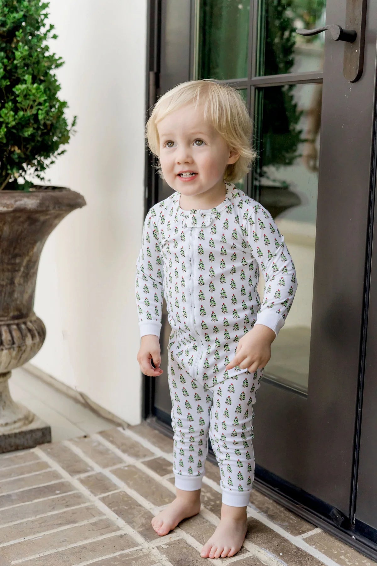 A toddler stands barefoot on a brick porch in front of a glass door, wearing Lila & Hayes Baby Parker Zipper Pajama made from soft Peruvian cotton with a green and yellow tree pattern.