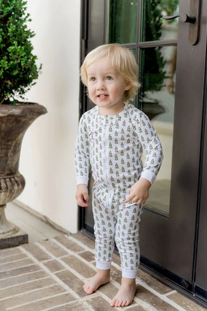A toddler stands barefoot on a brick porch in front of a glass door, wearing Lila & Hayes Baby Parker Zipper Pajama made from soft Peruvian cotton with a green and yellow tree pattern.