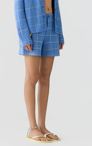A person wears the Guest in Residence Plaid Short with a matching blue plaid jacket, yellow bikini top, layered necklaces, and flat sandals, standing against a plain white background.