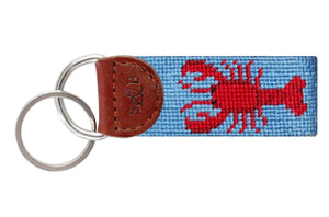 The Smathers & Branson Lobster Baby Blue Key Fob features a hand-stitched needlepoint strap, brown Italian leather end, sturdy metal keyring, and blue fabric with a red lobster design.