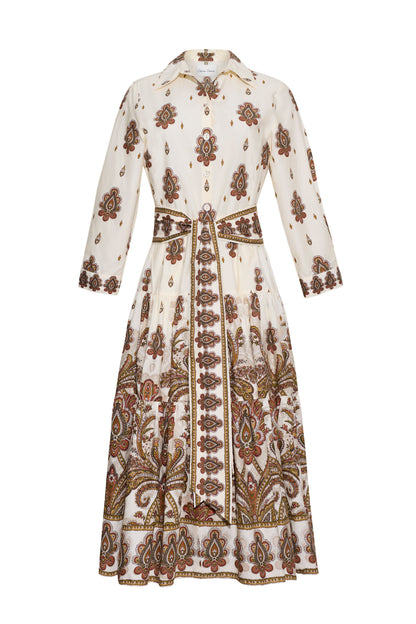 The Lolita Dress by Cara Cara is a long-sleeve, collared shirtdress in white, adorned with brown, rust, and olive paisley border print and floral patterns. It features a button-front closure, fitted waist, and a mid-length skirt.
