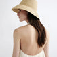 A woman with long brown hair wears the Janessa Leone Felix Hat and a backless cream dress, shown from behind against a plain light background.