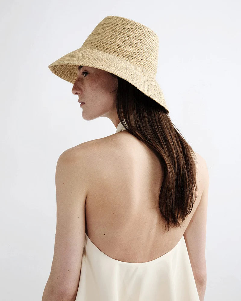 A woman with long brown hair wears the Janessa Leone Felix Hat and a backless cream dress, shown from behind against a plain light background.