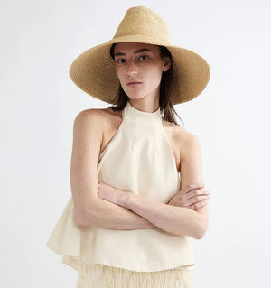 A woman stands with her arms crossed against a plain background, wearing the Janessa Leone Colette Hat and a sleeveless cream halter top.