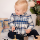 A toddler with blonde hair sits on the floor wearing Antoinette's Boys' Louis Blue Tartan Romper, gazing to the side. She is surrounded by toys and flowers, creating a charming holiday celebration scene.