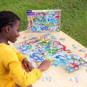 A girl in a yellow sweater assembles the Eeboo Love of Dragons 100-Piece Puzzle on a wooden table outdoors, with the dragon reference poster and the puzzle box nearby.