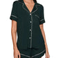 A woman wears the Eberjey Gisele Relaxed Short PJ Set in dark green, featuring a loose fit, short sleeves, white piping, and a chest pocket, standing against a plain beige background.