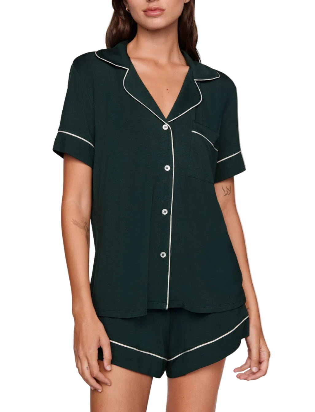 A woman wears the Eberjey Gisele Relaxed Short PJ Set in dark green, featuring a loose fit, short sleeves, white piping, and a chest pocket, standing against a plain beige background.