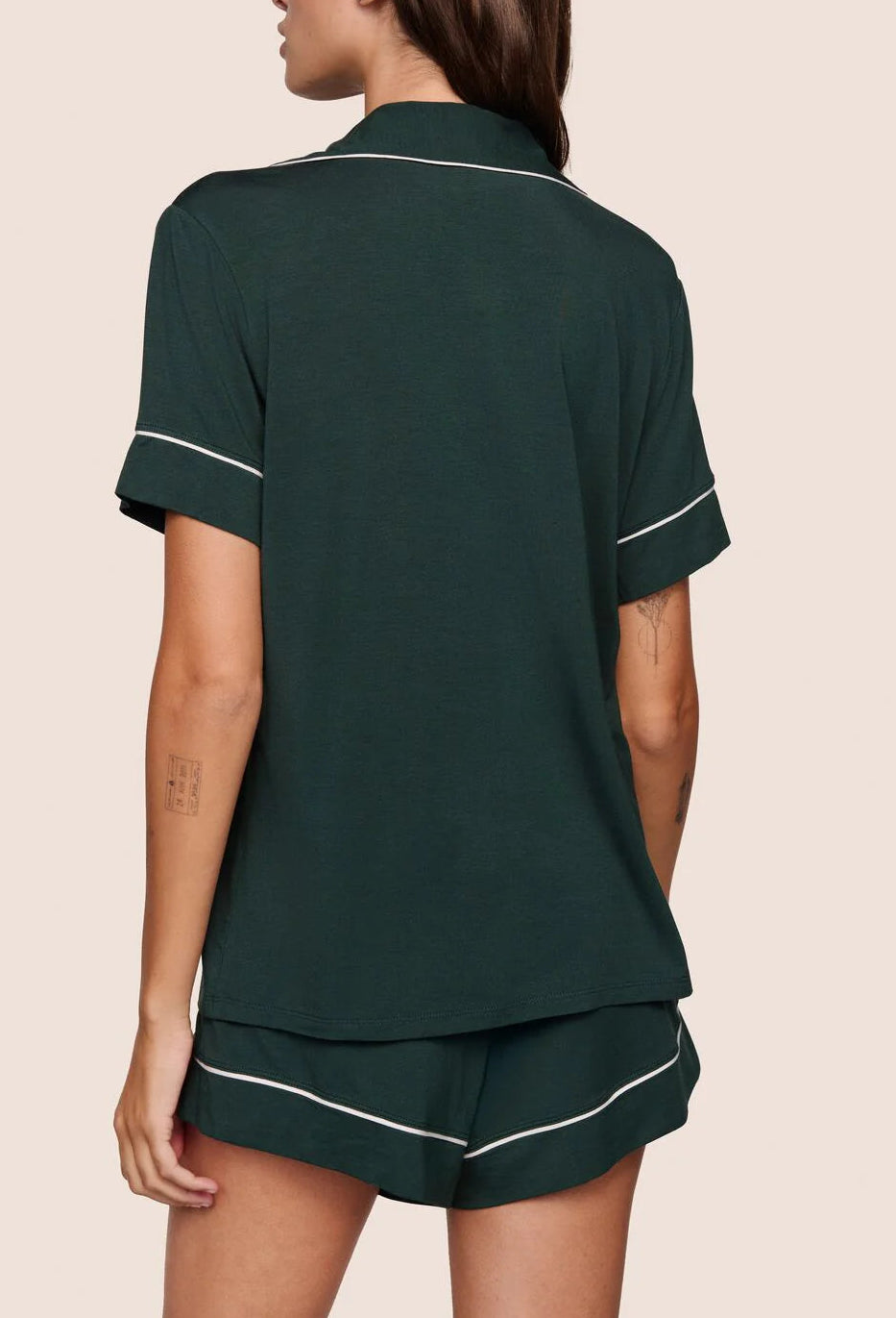 A person faces away, wearing the Eberjey Gisele Relaxed Short PJ Set—a dark green, short-sleeve pajama top and matching shorts with white trim—against a plain light background.