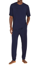 A person stands wearing the Eberjey Henry Long PJ Set from Eberjey, which includes a navy blue short-sleeve t-shirt and matching long pants made of a soft, breathable fabric with an elastic waistband for added comfort.