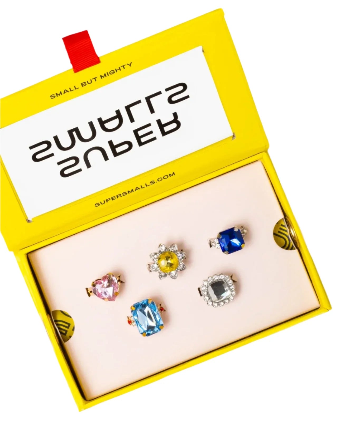 A yellow Super Smalls box labeled "SMALL BUT MIGHTY" holds the Power Lunch Ring Set—five colorful, gem-like rings displayed on a cream insert.