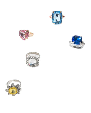 The Super Smalls Power Lunch Ring Set features five bold rings—a pink heart, blue rectangle, dark blue square, clear circle, and yellow oval—arranged on a white background for the perfect statement look.