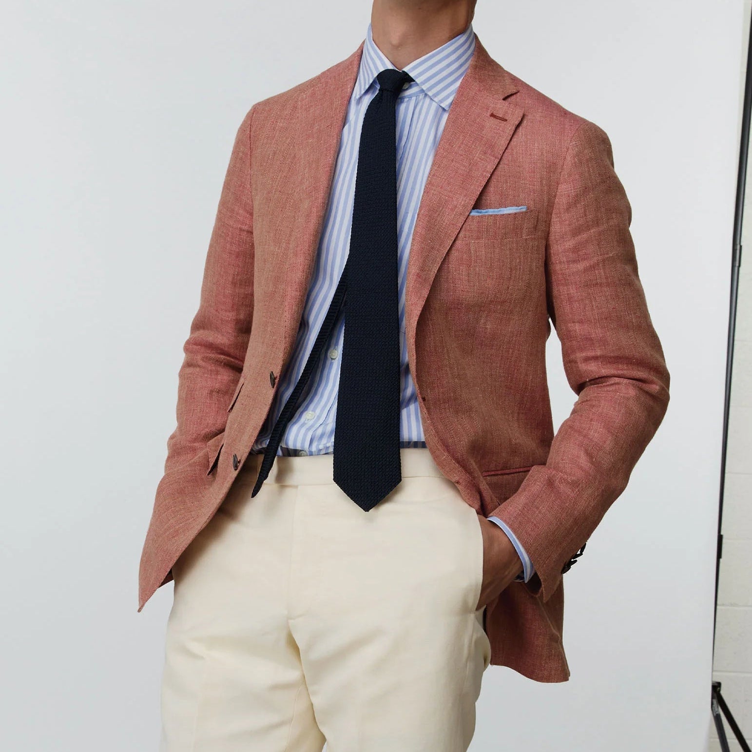 A person in the Sid Mashburn Kincaid No. 3 Jacket, paired with a light red, blue and white striped shirt, navy tie, and cream trousers stands with hands in pockets against a plain background.