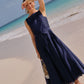 A woman wearing the Ann Mashburn Rose Dress—sleeveless, navy, pleated with a square neckline—stands on a sandy beach near the water, shielding her eyes from the sun with her hand.