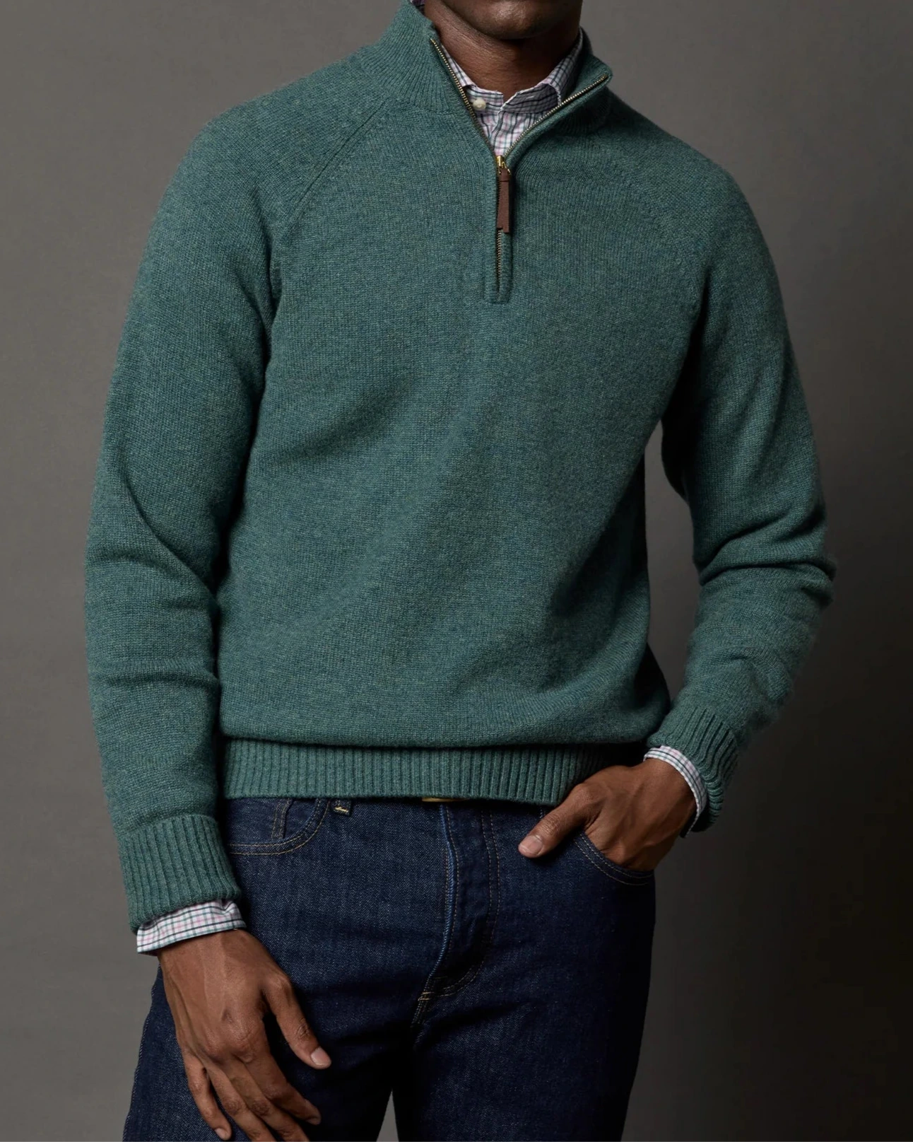 A person wears a Sid Mashburn Half-Zip Sweater in green over a plaid shirt with dark blue jeans, standing against a plain gray background.