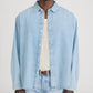 A person stands against a plain background wearing the Frame Classic Denim Shirt in light blue, designed in a relaxed fit over a white knit top. The outfit is completed with blue jeans and a brown belt. Crafted from 100% cotton, the Classic Denim Shirt offers superior comfort and breathability.