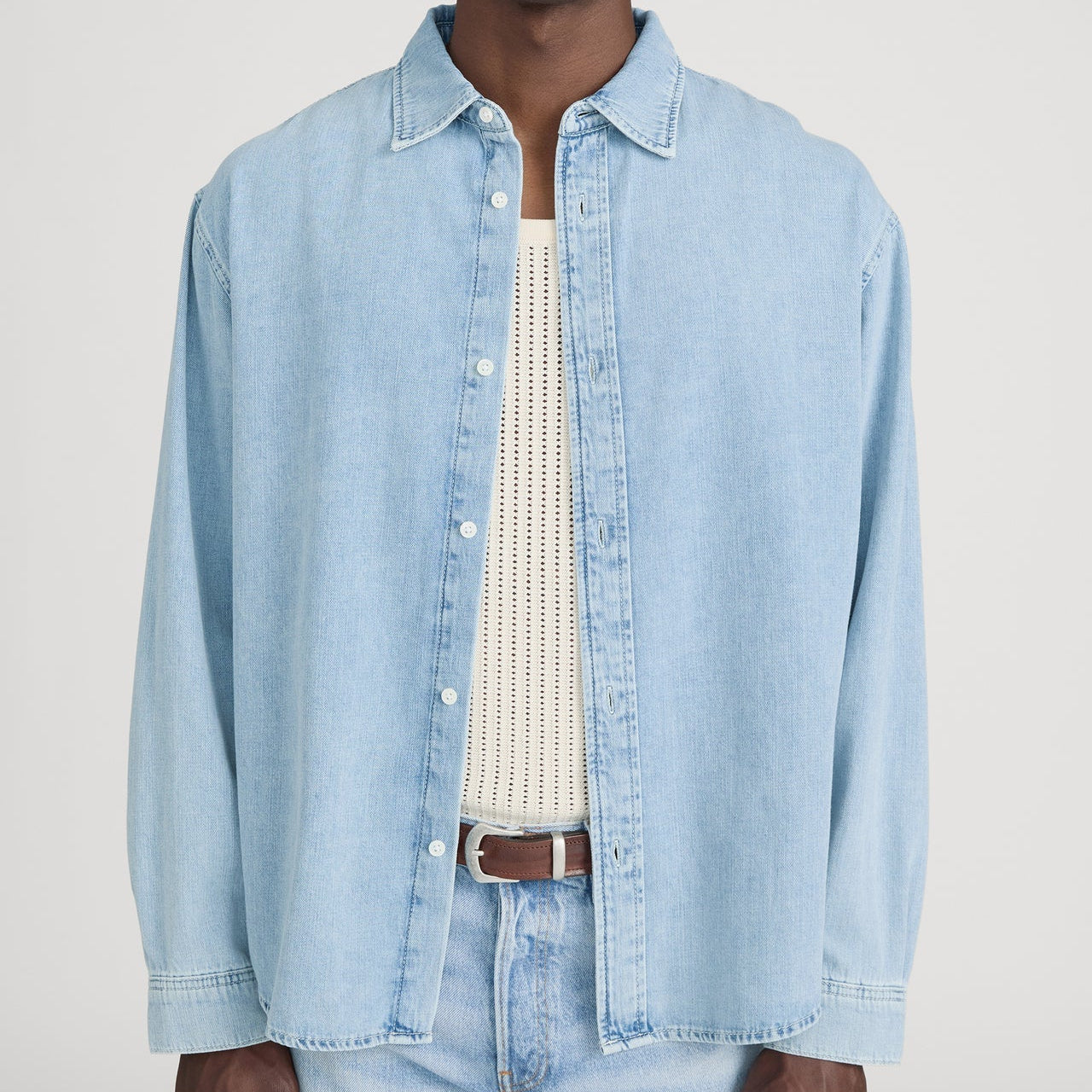 A person stands against a plain background wearing the Frame Classic Denim Shirt in light blue, designed in a relaxed fit over a white knit top. The outfit is completed with blue jeans and a brown belt. Crafted from 100% cotton, the Classic Denim Shirt offers superior comfort and breathability.