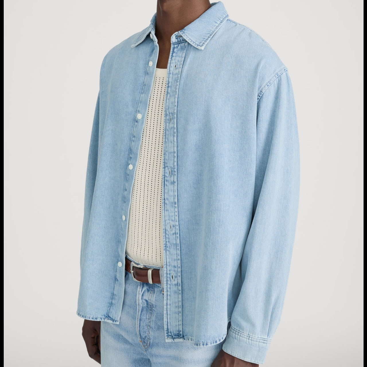 A person wearing the Frame Classic Denim Shirt—a relaxed fit, 100% cotton denim shirt—over a cream knit tank top, paired with light blue jeans and a brown belt.