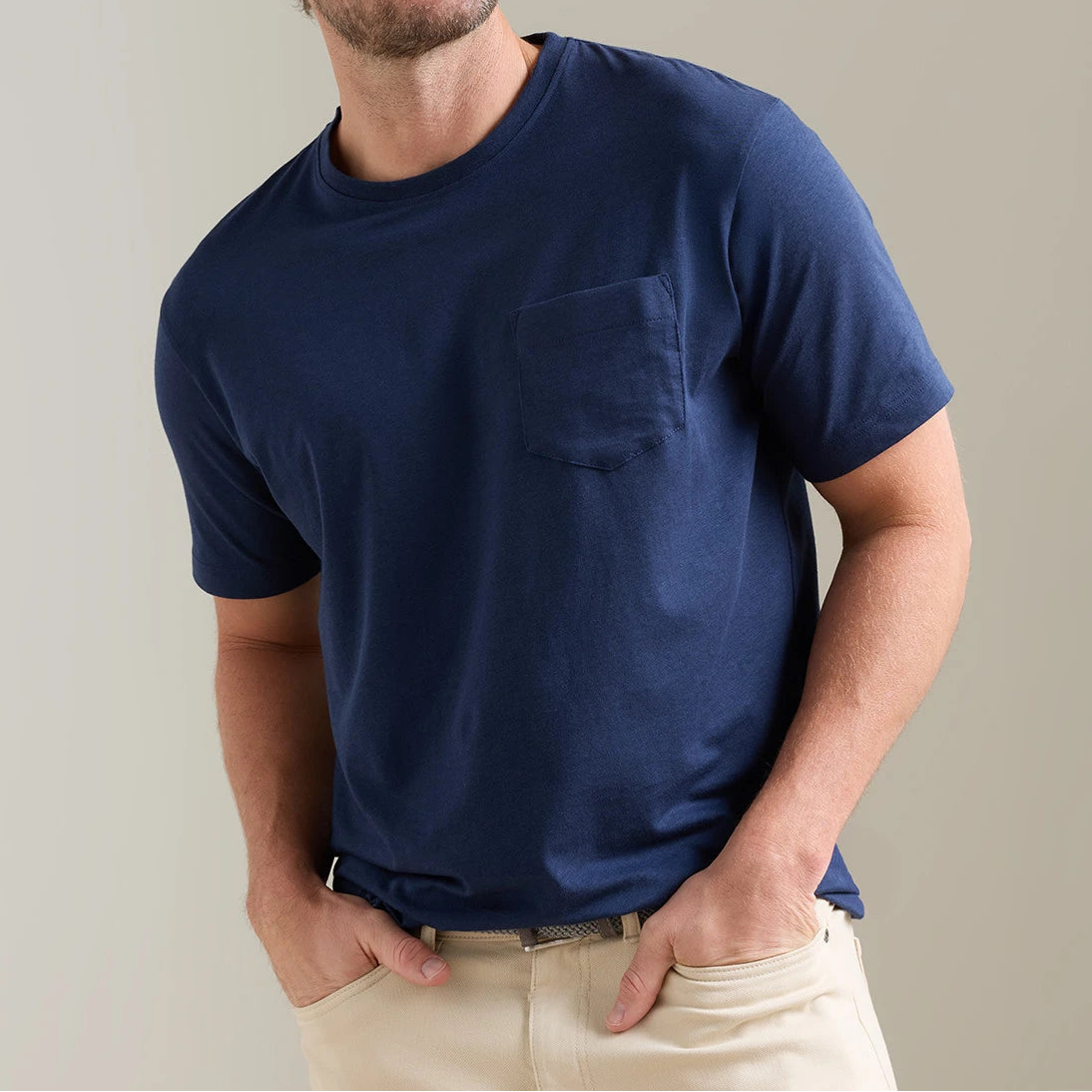 A man wearing the Peter Millar Crown Comfort Short-Sleeve Pocket Tee in navy blue paired with beige pants stands with his hands in his pockets against a plain light background.