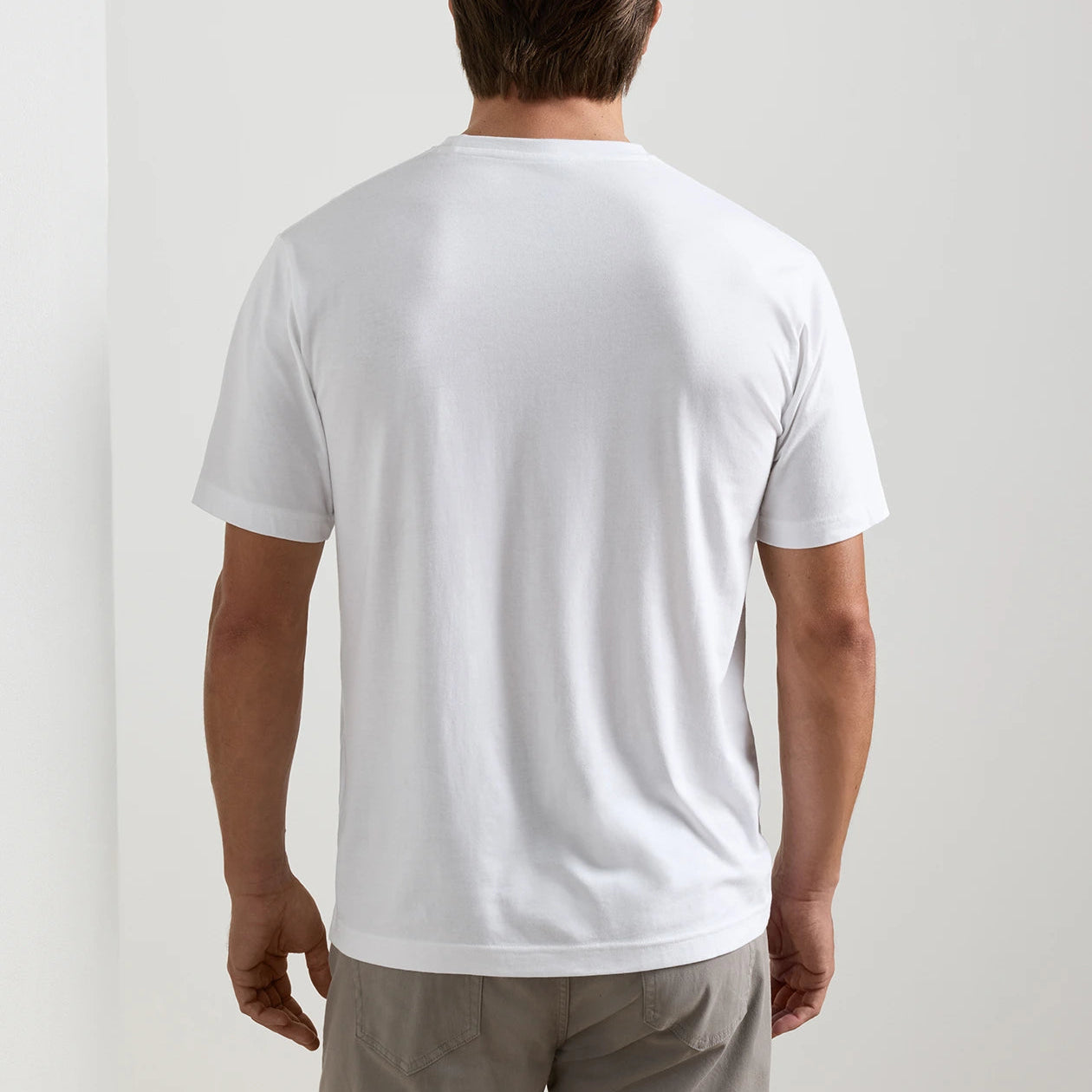 A man stands facing away from the camera, wearing the Peter Millar Crown Comfort Short-Sleeve Pocket Tee, crafted from soft organic cotton, paired with light gray pants against a plain white background.