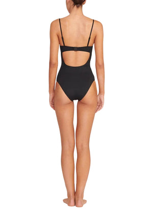 A woman stands barefoot facing away, wearing the Staud Maeve One Piece swimsuit featuring a black bustier bodice, adjustable shoulder straps, and a back cut-out detail.