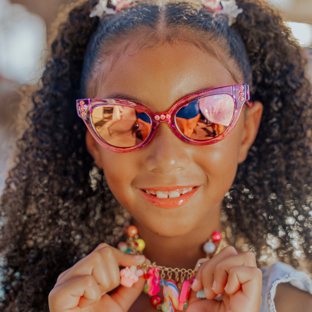 A young girl with curly hair beams outdoors in bright sunlight, wearing Bling2O’s Girls’ Malibu Beach Sunglasses and a cheerful beaded necklace. These stylish sunglasses feature UV-protected lenses, making them perfect for sunny days.
