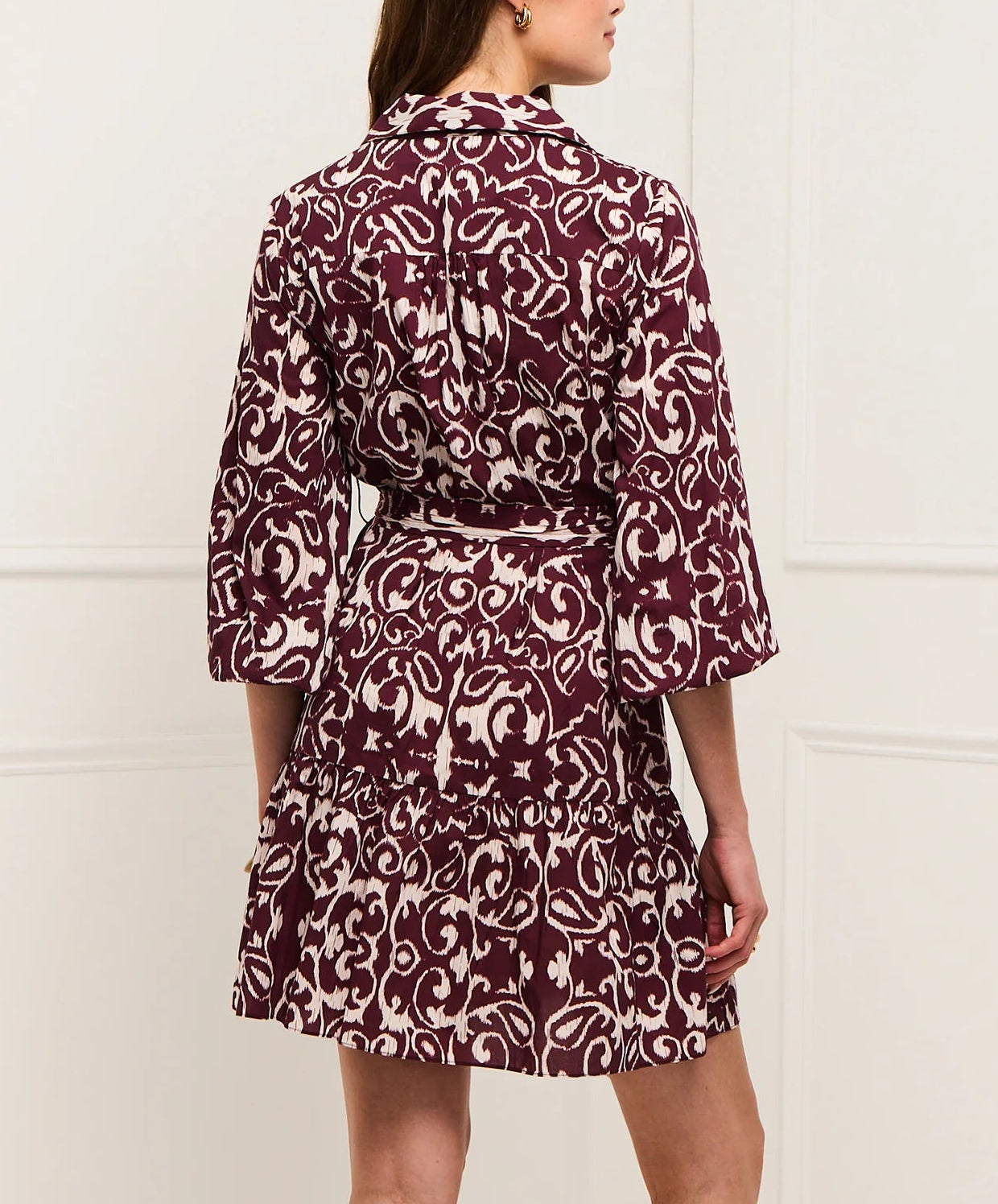 A woman faces away wearing the Figue Marcy Dress, featuring a maroon and white pattern in cotton, three-quarter sleeves, and a gracefully flared, tiered skirt.
