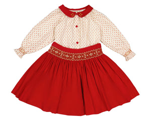 The Antoinette Girls' Marie Red Floral Smocked Skirt and Blouse Set features a cream long-sleeved handmade blouse with a red collar, red buttons, and a bright red corduroy skirt with floral embroidery at the waist.