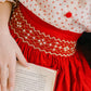 A child wears the Antoinette Girls' Marie Red Floral Smocked Skirt and Blouse Set while holding an open book, revealing part of its text.