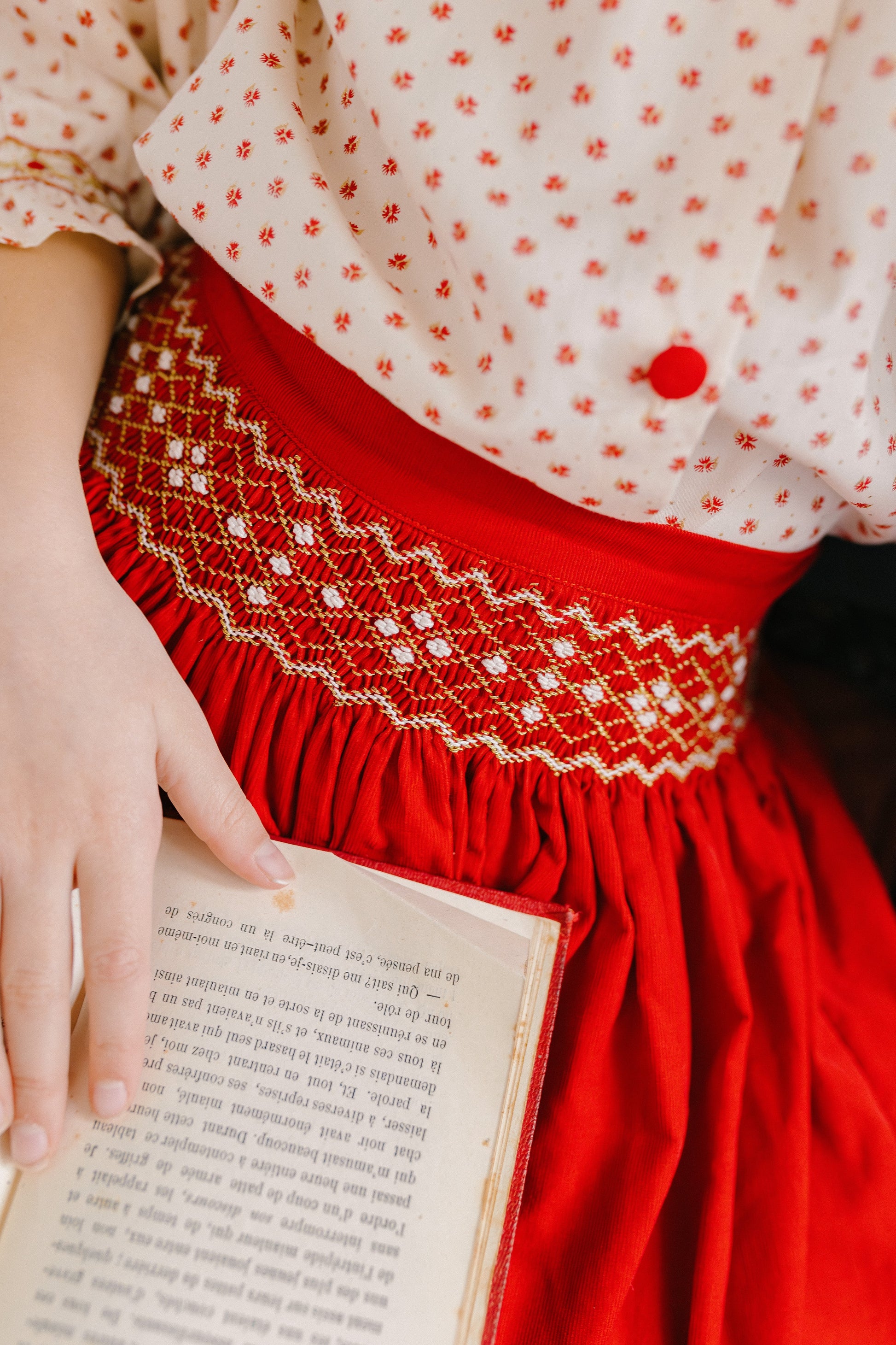 A child wears the Antoinette Girls' Marie Red Floral Smocked Skirt and Blouse Set while holding an open book, revealing part of its text.