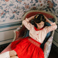 A young girl wears the Antoinette Girls' Marie Red Floral Smocked Skirt and Blouse Set, sitting on a pink chaise lounge against floral wallpaper, looking down with her hands behind her head.