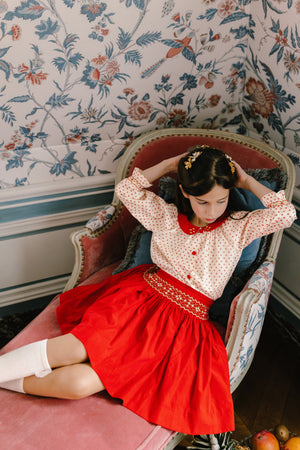 A young girl wears the Antoinette Girls' Marie Red Floral Smocked Skirt and Blouse Set, sitting on a pink chaise lounge against floral wallpaper, looking down with her hands behind her head.