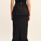 A woman faces away, wearing a sleeveless black top with a back buckle and the POSSE Matilda Column Skirt—a premium linen piece with a high-rise waist, ruffled hem, back slit—paired with black sandals.