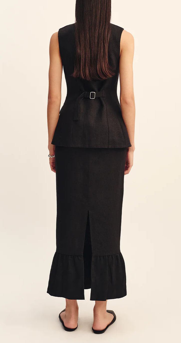 A woman faces away, wearing a sleeveless black top with a back buckle and the POSSE Matilda Column Skirt—a premium linen piece with a high-rise waist, ruffled hem, back slit—paired with black sandals.