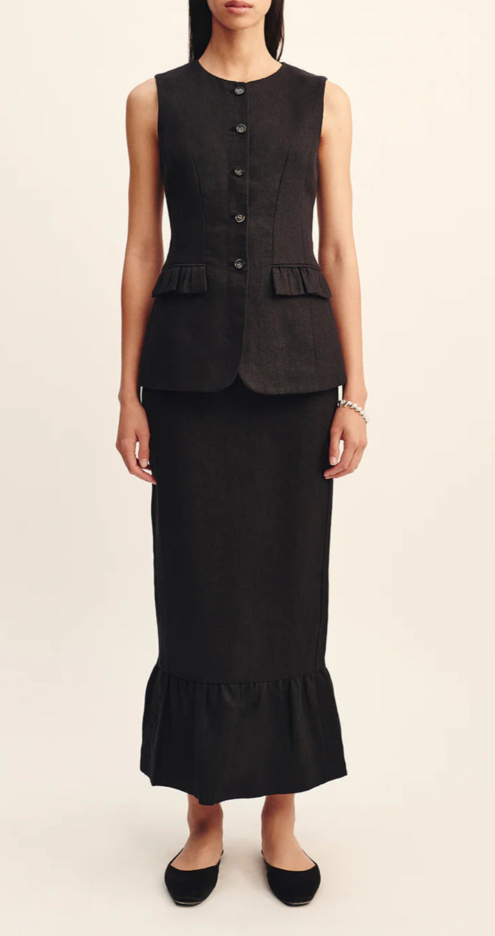 A woman wears the POSSE Matilda Column Skirt—a high-rise, ruffled-hem piece—paired with a sleeveless, button-up black vest with ruffle pockets and black flats.