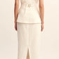 A woman faces away, wearing a sleeveless cream top with a belt and the POSSE Matilda Column Skirt in premium linen, featuring a high-rise waist, back slit, and ruffle hem. She finishes the look with black sandals.