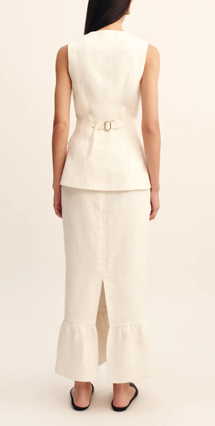 A woman faces away, wearing a sleeveless cream top with a belt and the POSSE Matilda Column Skirt in premium linen, featuring a high-rise waist, back slit, and ruffle hem. She finishes the look with black sandals.