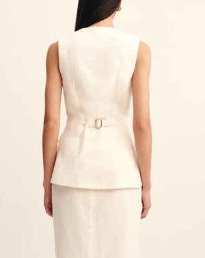 A woman is seen from the back wearing the POSSE Matilda Gilet, a sleeveless off-white linen piece with a tailored fit and waist buckle, styled with a matching skirt.