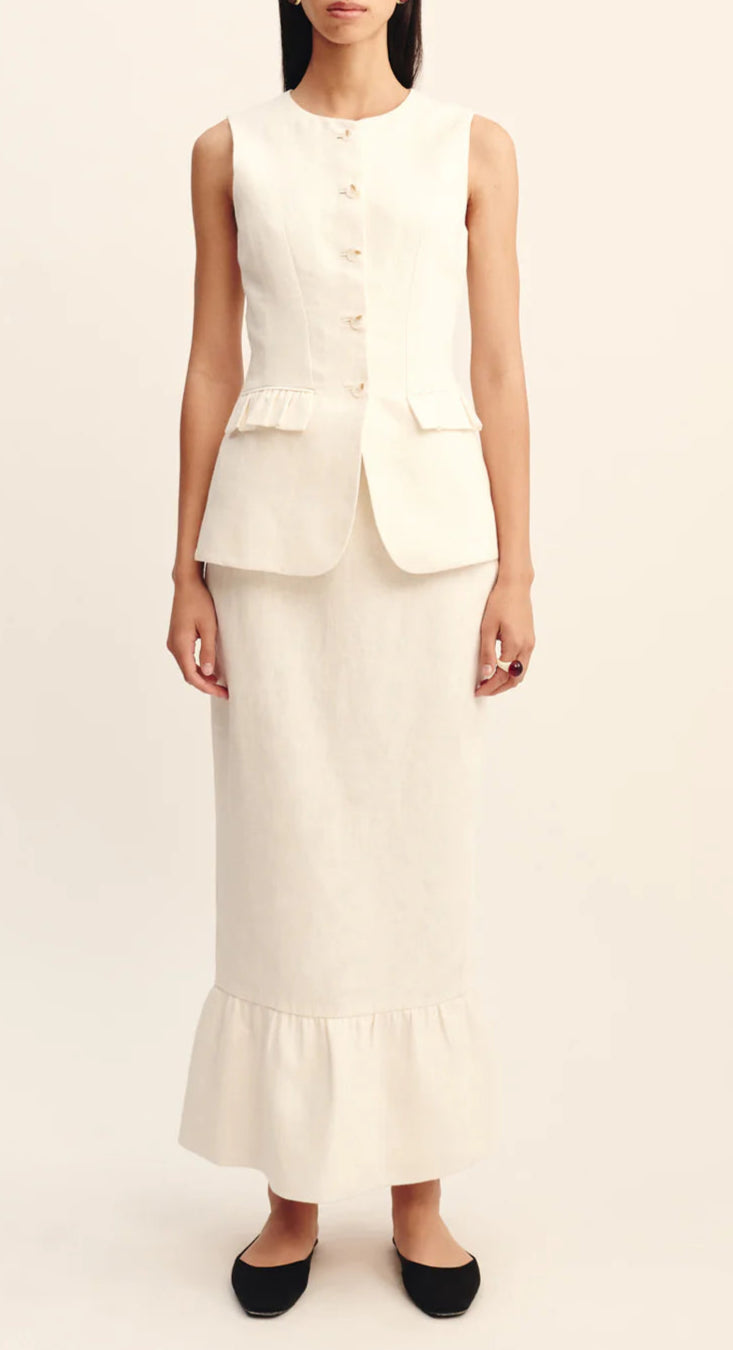 A woman wears a sleeveless, buttoned ivory vest with peplum detail and the POSSE Matilda Column Skirt in premium linen, featuring a high-rise waist and ruffled hem, paired with black flats.