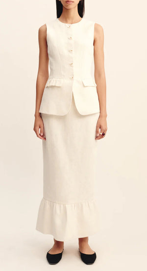 A woman wears a sleeveless, buttoned ivory vest with peplum detail and the POSSE Matilda Column Skirt in premium linen, featuring a high-rise waist and ruffled hem, paired with black flats.