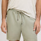 A person is wearing a light beige t-shirt and Onia's Air Linen Pull-On Short in sage green, featuring a stretch waist, shown from shoulders to mid-thigh.