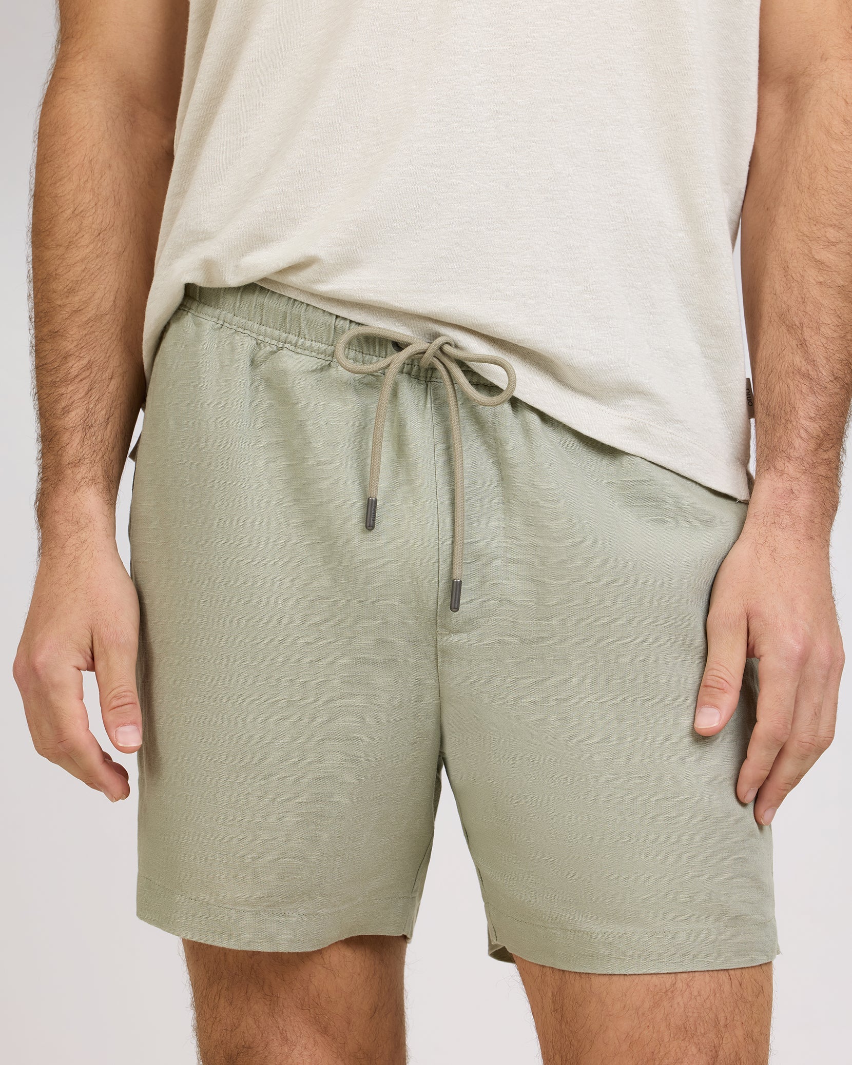 A person is wearing a light beige t-shirt and Onia's Air Linen Pull-On Short in sage green, featuring a stretch waist, shown from shoulders to mid-thigh.
