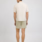 A man, facing away from the camera, wears a light short-sleeve shirt and beige Onia Land to Water Short 6" with quick-dry, four-way stretch fabric, styled with brown loafers against a plain background.