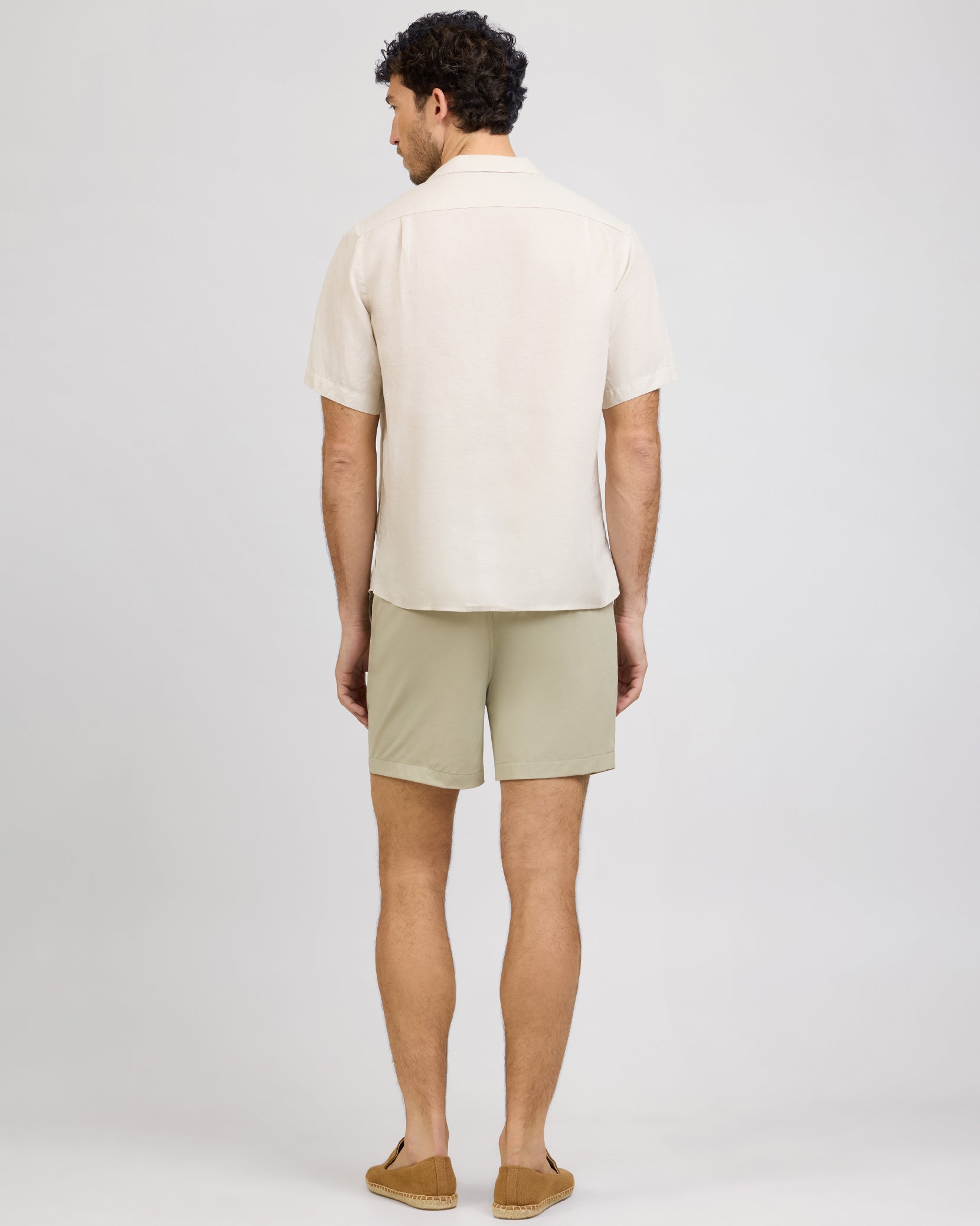 A man, facing away from the camera, wears a light short-sleeve shirt and beige Onia Land to Water Short 6" with quick-dry, four-way stretch fabric, styled with brown loafers against a plain background.