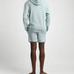 Person stands with back to camera wearing a light blue hoodie, Onia Slub Terry Short, and brown slide sandals against a plain background.
